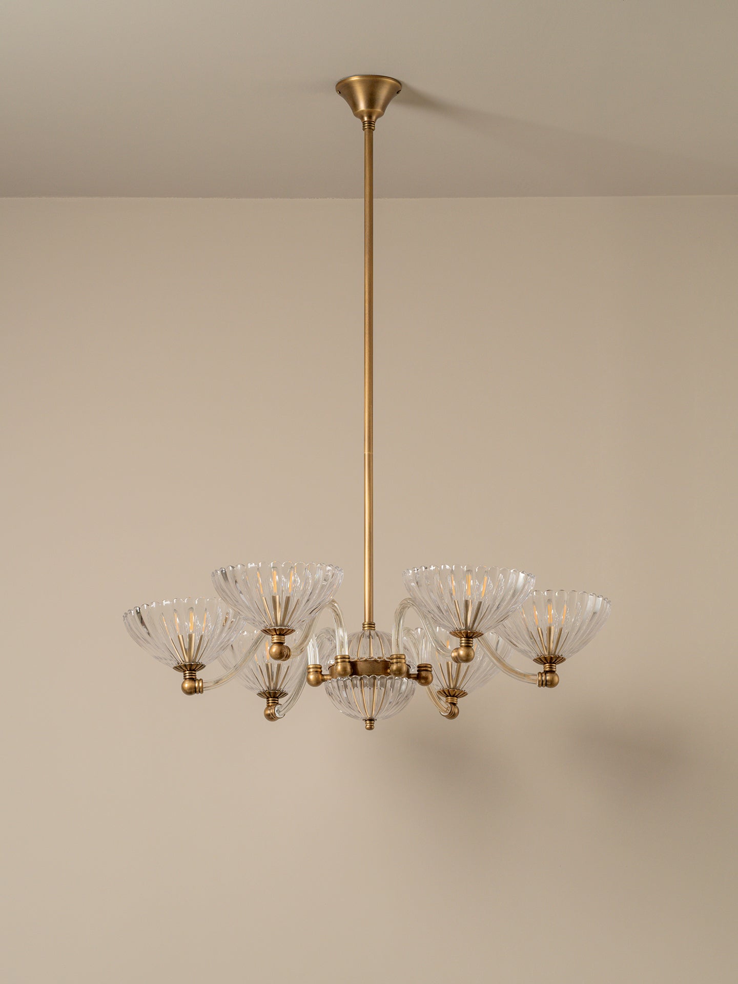 A modern six-light chandelier with a brushed brass or gold-finished metal frame and clear ribbed glass shades, suspended from a matching ceiling canopy against a plain beige wall.