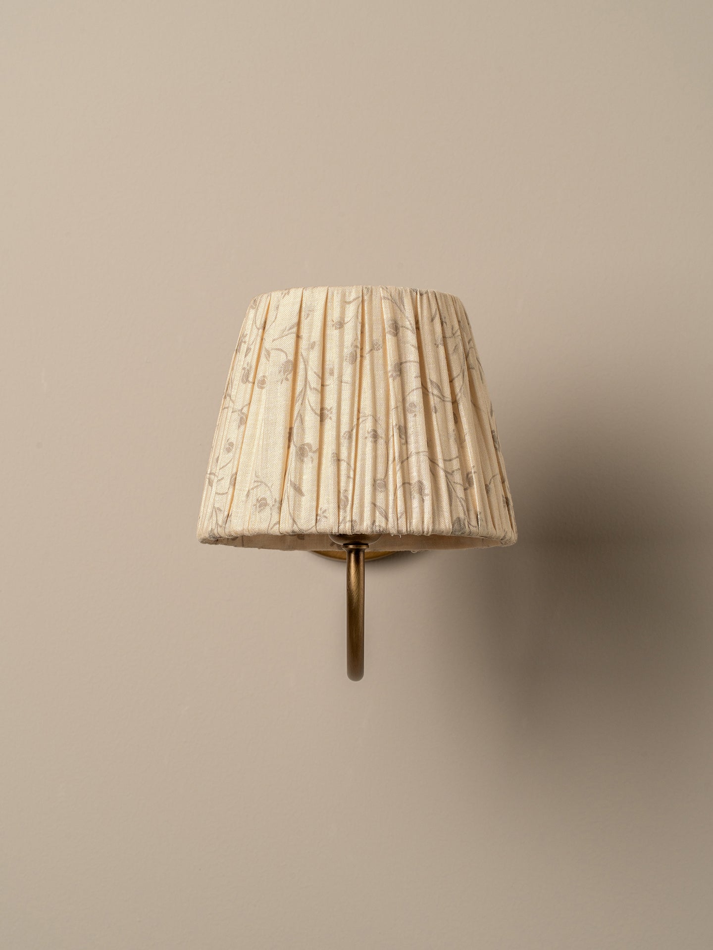 A vintage-style wall sconce featuring a pleated fabric shade in cream with a delicate brown floral pattern, mounted on a brass-colored metal bracket against a solid beige wall.