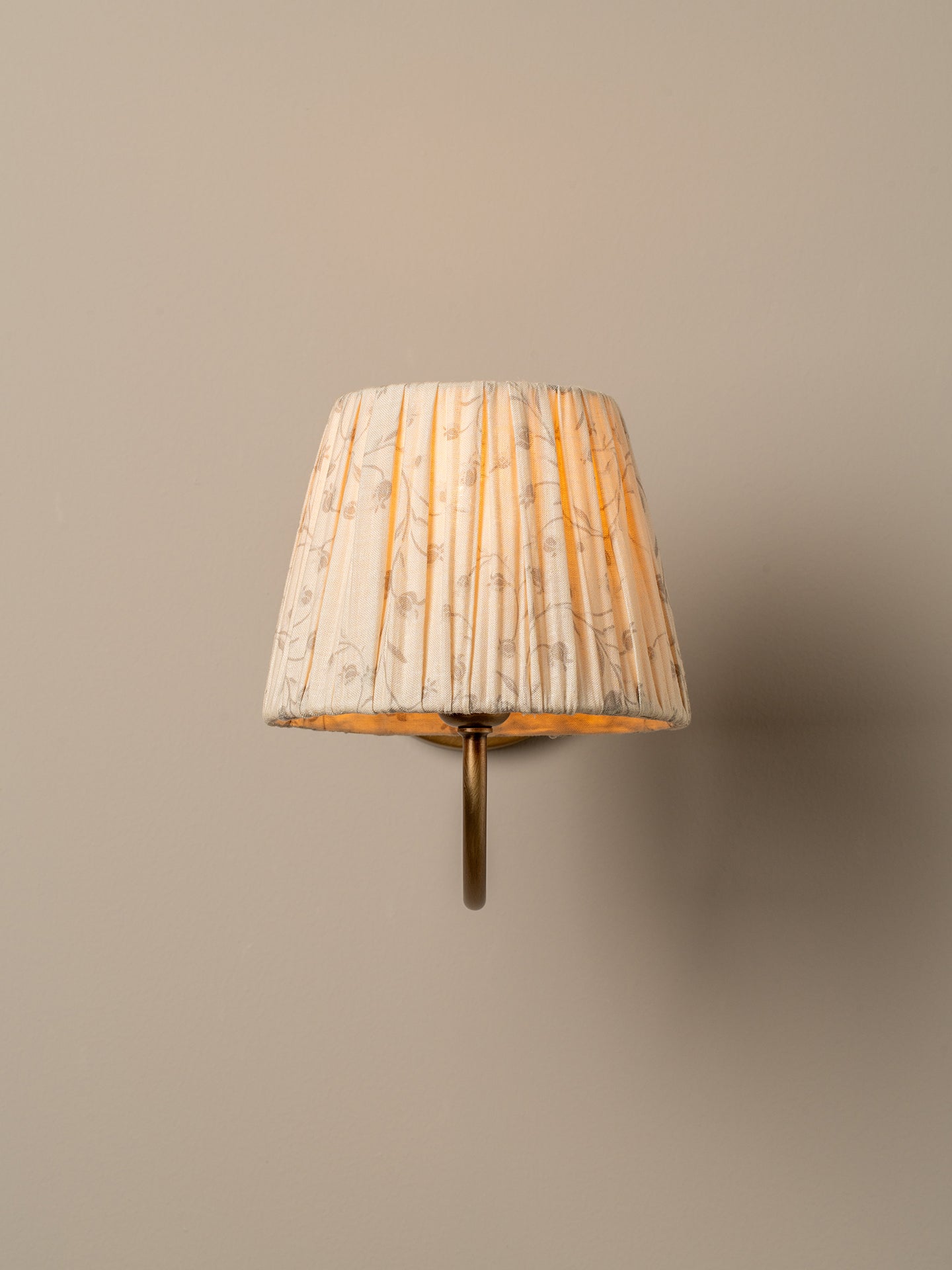A wall-mounted sconce with a pleated fabric shade in a warm cream and beige floral pattern, illuminated with a soft yellow glow, mounted on a solid beige wall.