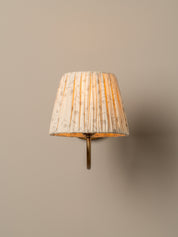 A wall-mounted sconce with a pleated fabric shade in a warm cream and beige floral pattern, illuminated with a soft yellow glow, mounted on a solid beige wall.