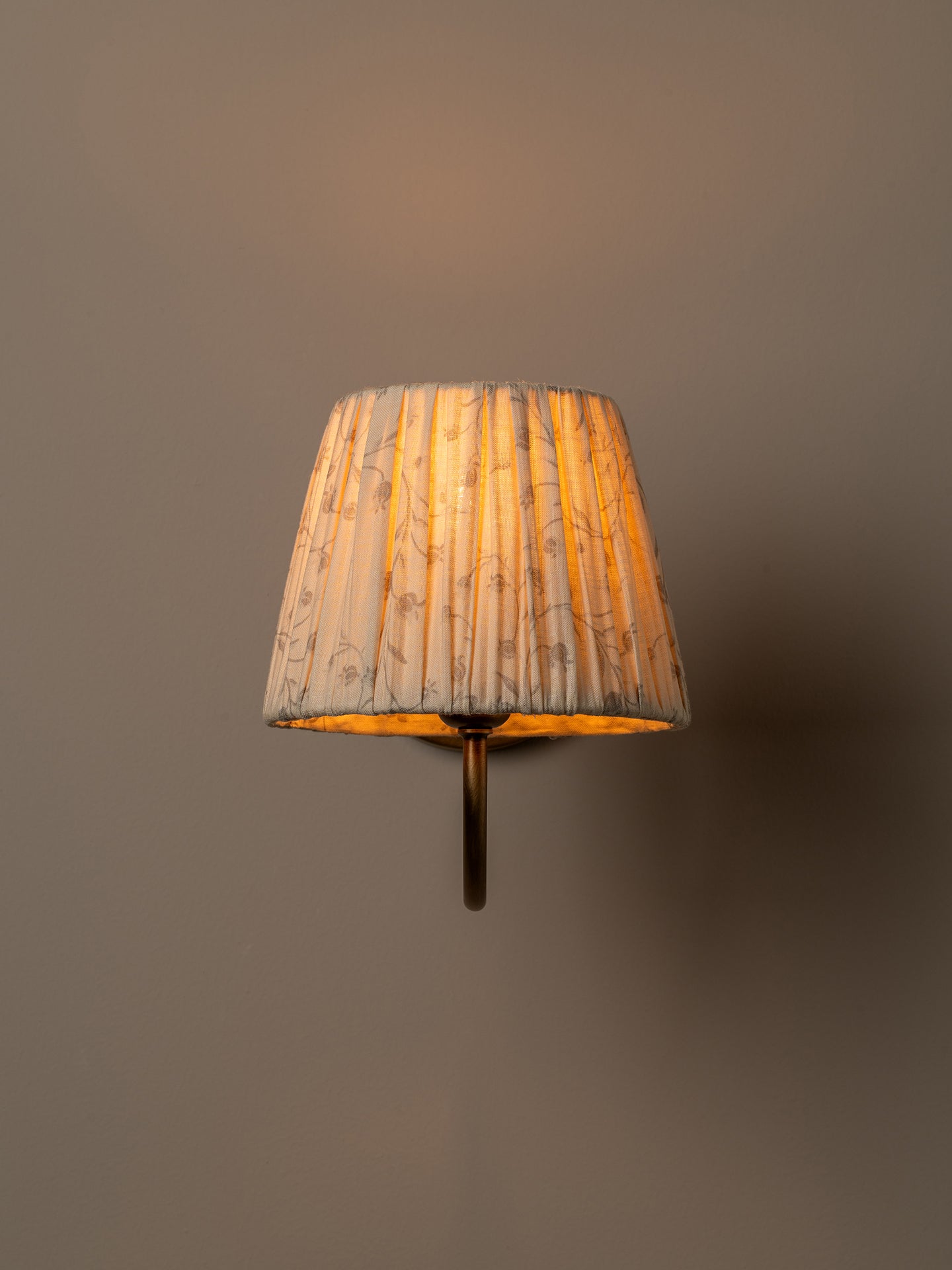 A wall-mounted sconce with a pleated fabric shade in a warm beige and brown floral pattern, glowing with a soft golden light against a solid taupe-gray background.