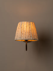A wall-mounted sconce with a pleated fabric shade in a warm beige and brown floral pattern, glowing with a soft golden light against a solid taupe-gray background.