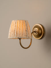 A wall-mounted sconce with a pleated fabric shade in a beige and white floral pattern, illuminated with a warm yellow glow, attached to an antique brass curved arm and circular wall plate against a neutral beige wall.