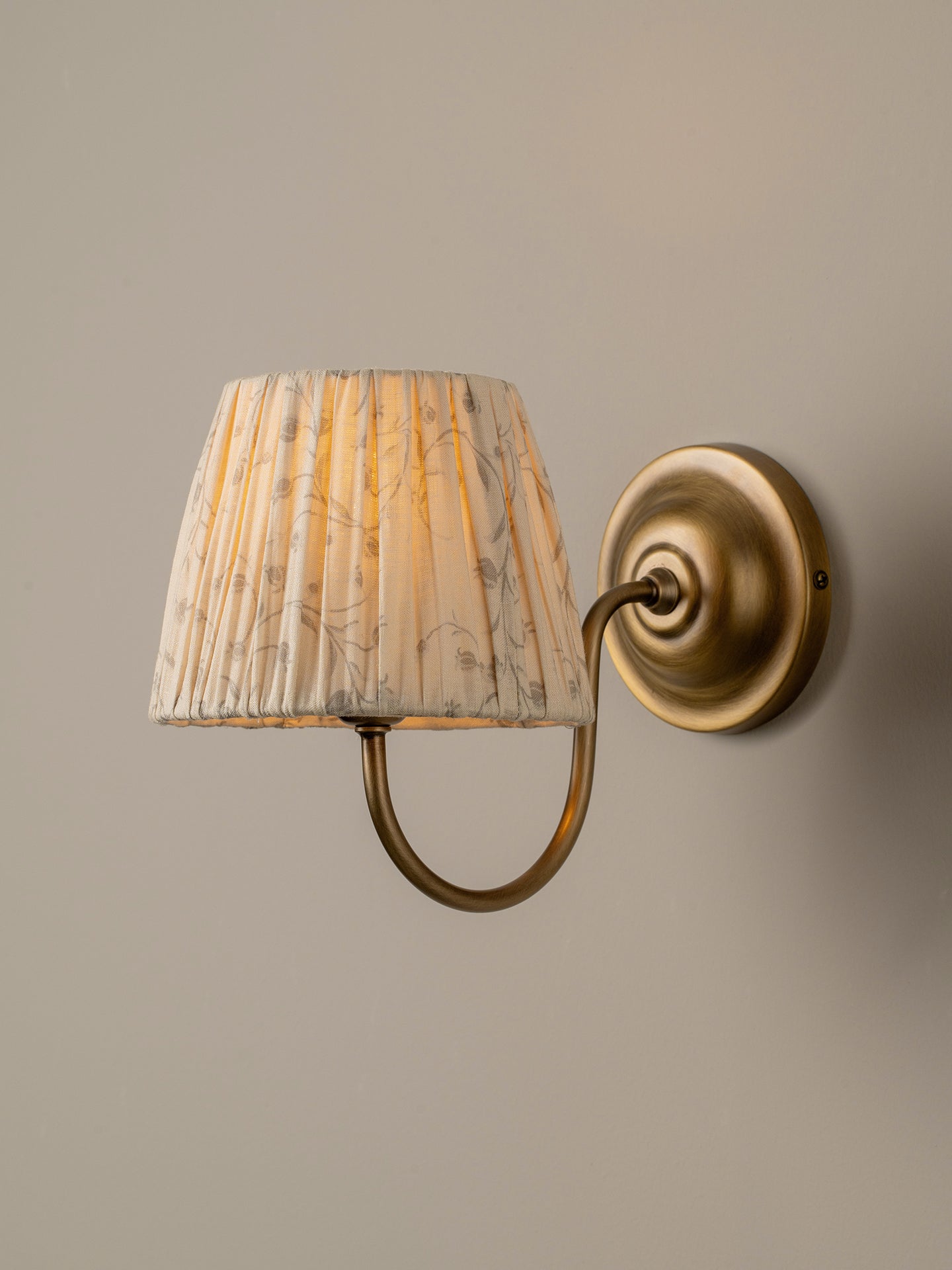 A wall-mounted sconce with a pleated fabric shade featuring a gray and white marble pattern, illuminated with a warm yellow glow, attached to an antique brass curved arm and circular wall plate against a neutral beige wall.