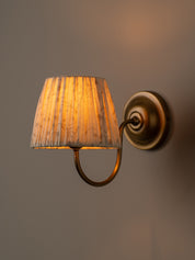 A wall-mounted sconce with a pleated fabric shade in a warm cream and brown pattern, illuminated with a golden glow, attached to an antique bronze metal arm against a neutral gray wall.