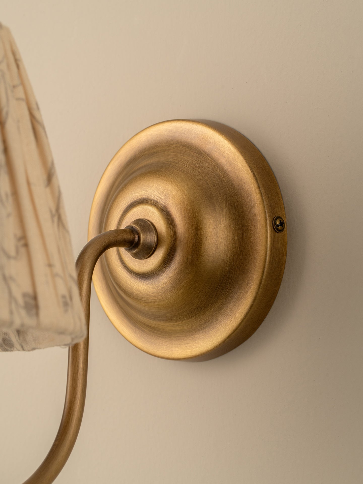 Close-up of a wall-mounted light fixture with a round brushed brass backplate and curved arm, shown against a warm beige wall with part of a patterned lampshade visible.