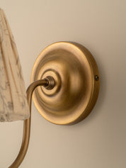 Close-up of a wall-mounted light fixture with a round brushed brass backplate and curved arm, shown against a warm beige wall with part of a patterned lampshade visible.
