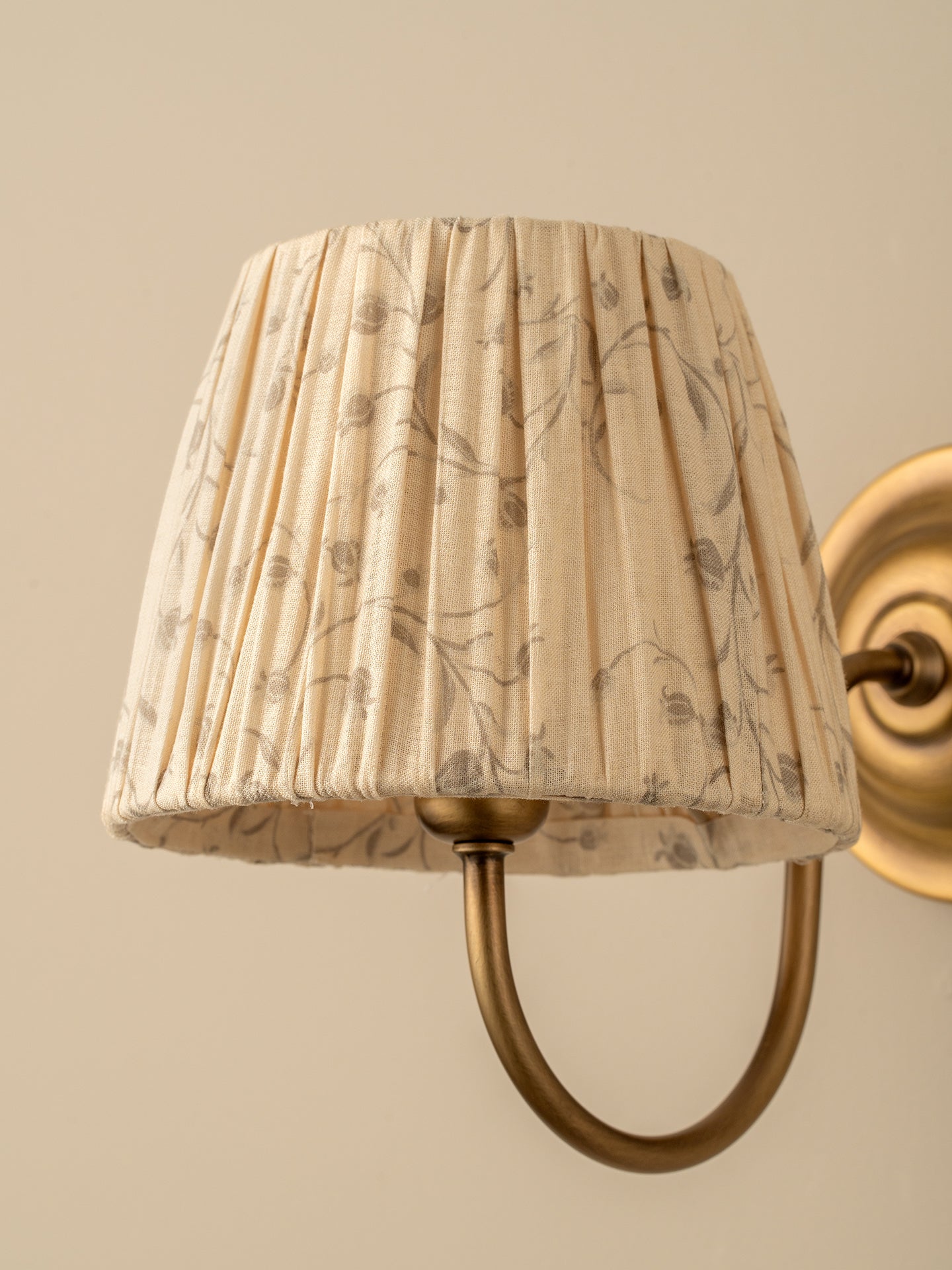 A close-up of a pleated fabric wall lampshade with a beige and brown botanical pattern, mounted on a brass U-shaped wall sconce against a warm beige wall.