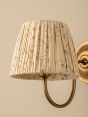 A close-up of a pleated fabric wall lampshade with a beige and brown botanical pattern, mounted on a brass U-shaped wall sconce against a warm beige wall.