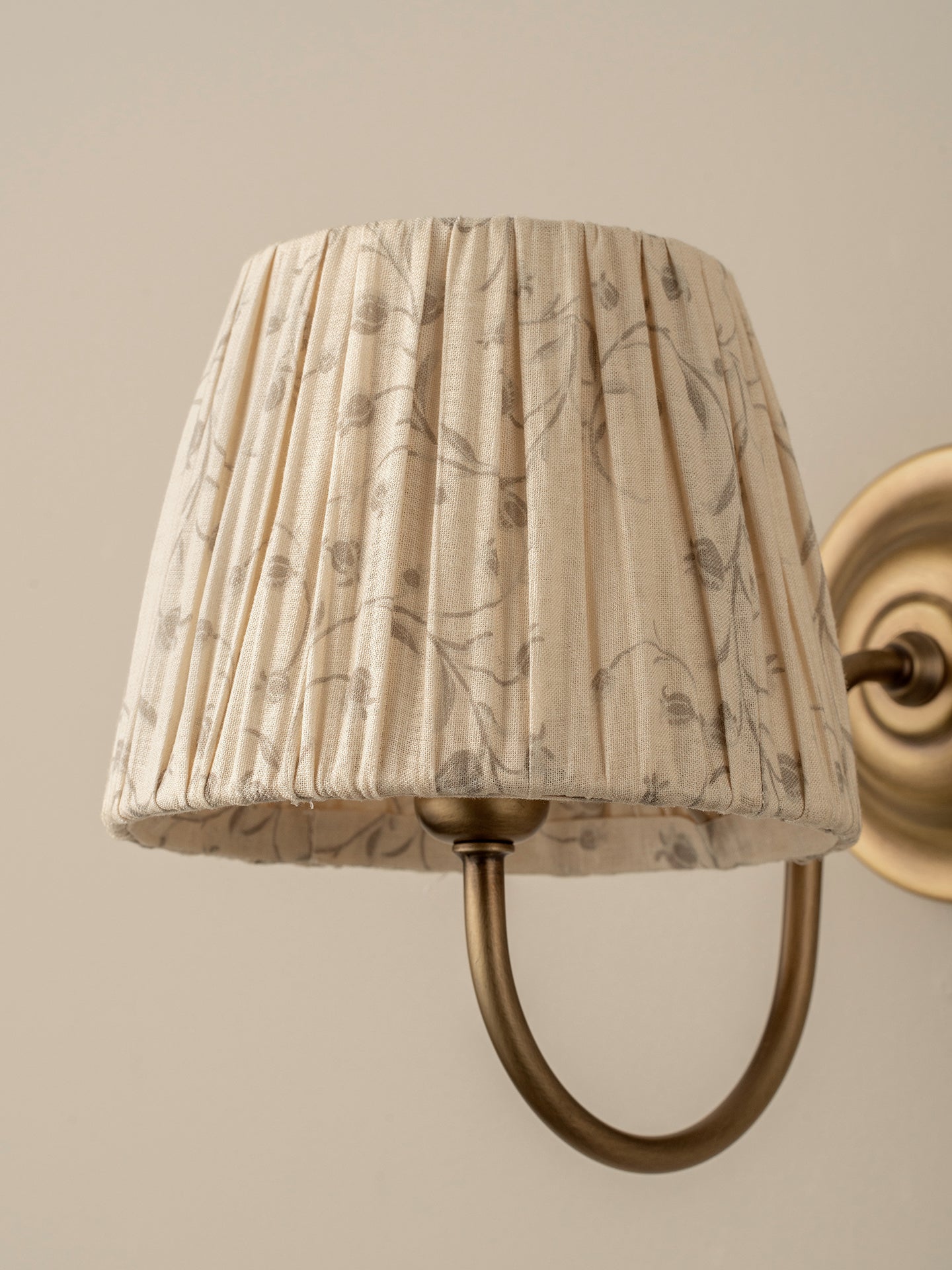 A close-up of a pleated fabric lampshade with a delicate gray botanical pattern, mounted on a warm brass U-shaped wall sconce against a neutral beige wall.