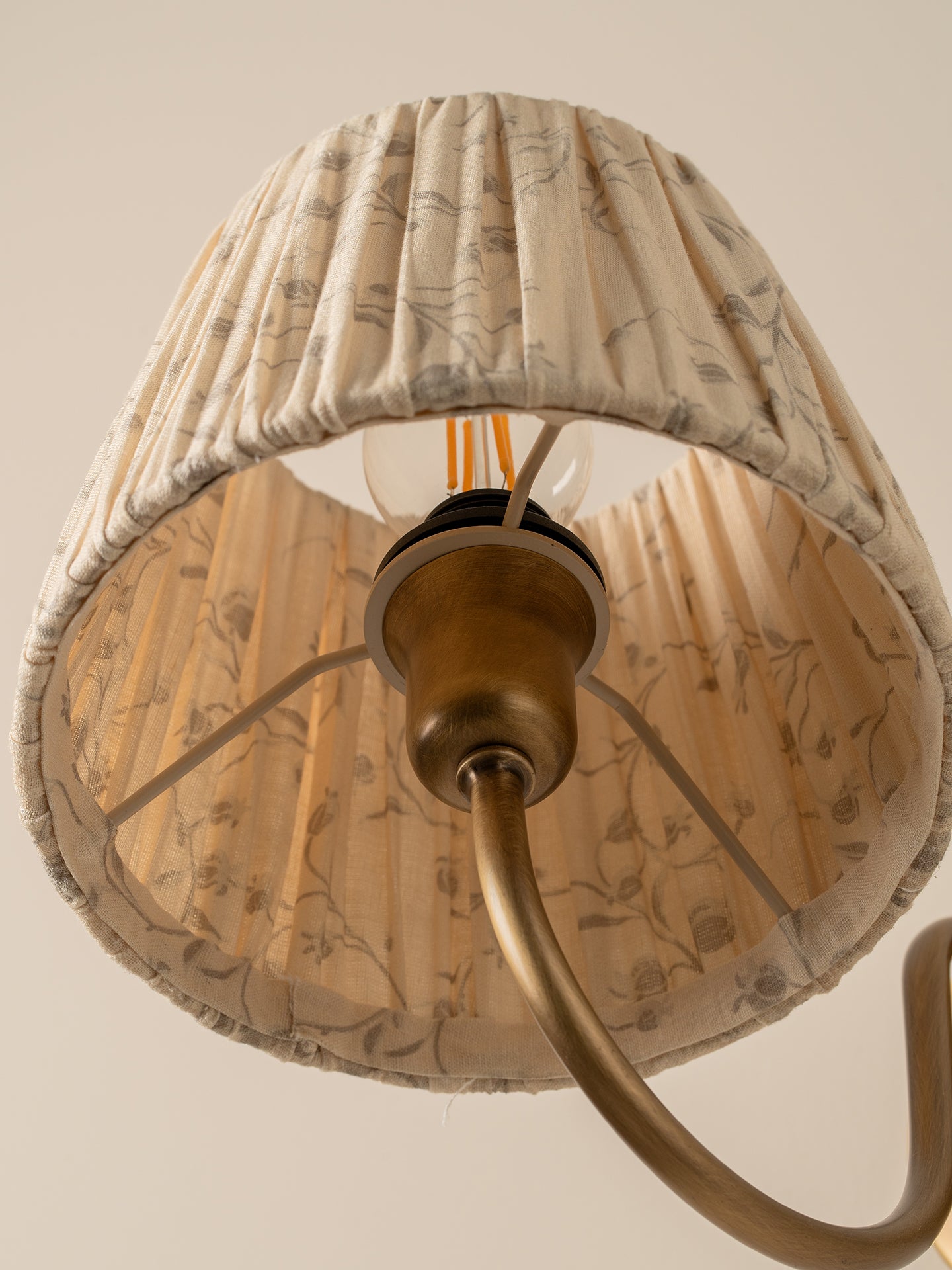 A close-up of a pleated fabric lampshade with a beige and brown botanical pattern, hanging from a brass-colored ceiling fixture against a plain beige wall.