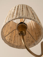 A close-up of a pleated fabric lampshade with a beige and brown botanical pattern, hanging from a brass-colored ceiling fixture against a plain beige wall.
