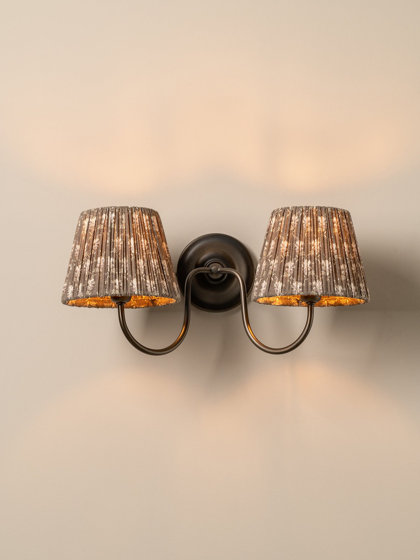A two-light wall sconce with textured metallic silver-gray fabric shades and warm gold-toned interiors, mounted on a dark metal wall plate against a warm beige wall.