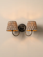 A two-light wall sconce with textured metallic silver-gray fabric shades and warm gold-toned interiors, mounted on a dark metal wall plate against a warm beige wall.