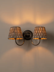 A two-light wall sconce with dark metal arms and textured gray fabric shades, shown lit against a warm beige wall.