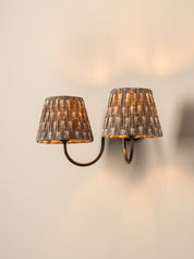 A two-light wall sconce with textured, pleated drum shades in a distressed brown and gold finish, mounted on a warm beige wall.