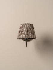A wall-mounted sconce with a pleated fabric shade in a distressed gray and white pattern, mounted on a dark metal bracket against a plain beige wall.