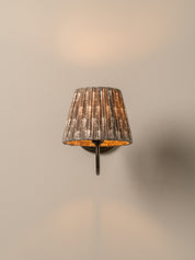 A wall-mounted sconce with a pleated fabric shade in a distressed gray and white pattern, mounted on a dark metal bracket against a plain beige wall.