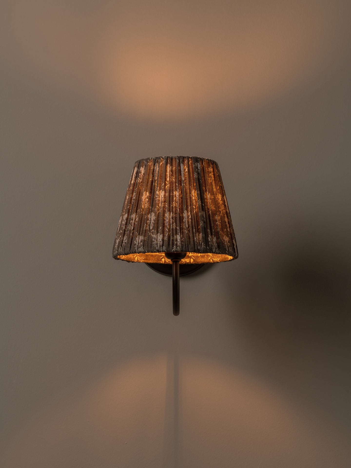 A wall-mounted sconce with a dark brown textured pleated fabric shade and a warm gold interior, shown lit against a neutral gray wall.