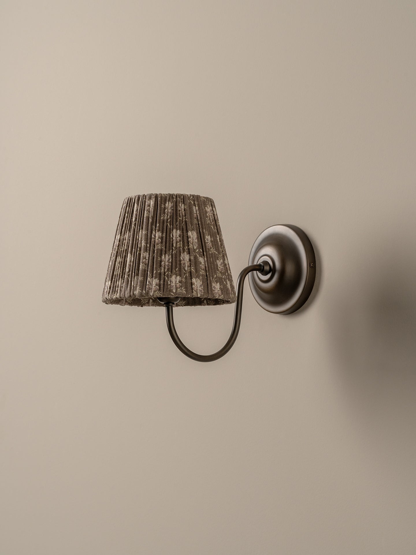 A wall-mounted sconce with a textured, pleated drum shade in a brown and beige pattern, attached to a dark metal curved arm and circular wall plate against a warm beige wall.