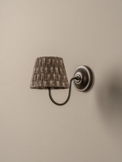 A wall-mounted sconce with a textured, pleated drum shade in a brown and beige pattern, attached to a dark metal curved arm and circular wall plate against a warm beige wall.