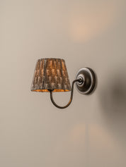 A wall-mounted sconce with a textured, distressed brown and gold pleated fabric shade and a dark metal curved arm, shown lit against a warm beige wall.