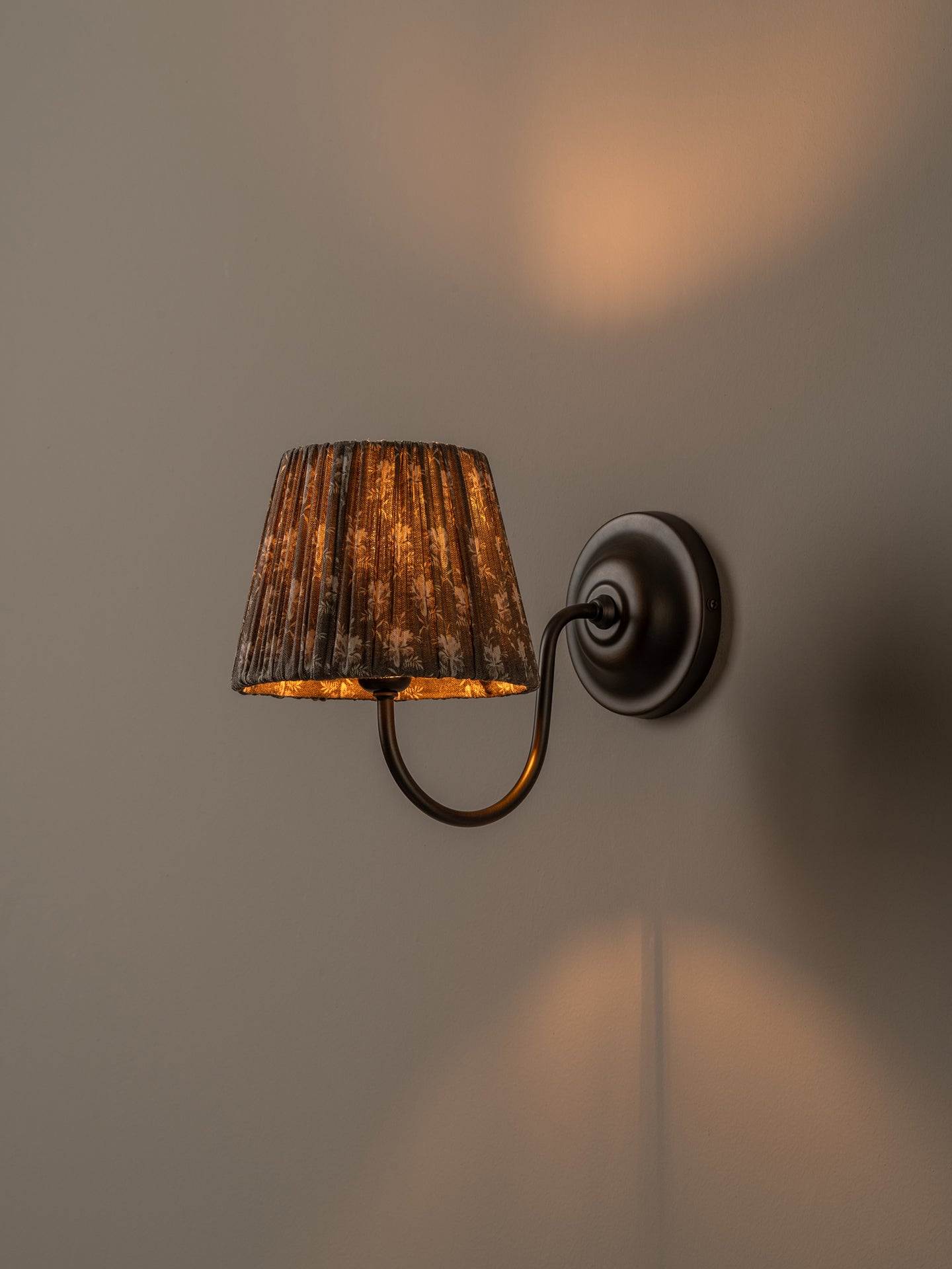 A wall-mounted sconce with a dark metal curved arm and circular backplate, featuring a textured brown fabric drum shade that glows warm yellow when lit, mounted on a plain beige wall.