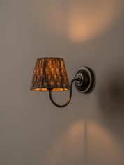 A wall-mounted sconce with a dark metal curved arm and circular backplate, featuring a textured brown fabric drum shade that glows warm yellow when lit, mounted on a plain beige wall.