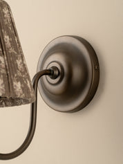 Close-up of a wall-mounted light fixture with a round dark bronze or oil-rubbed metal wall plate and curved arm, shown against a warm beige wall. Part of a patterned fabric shade is visible on the left.