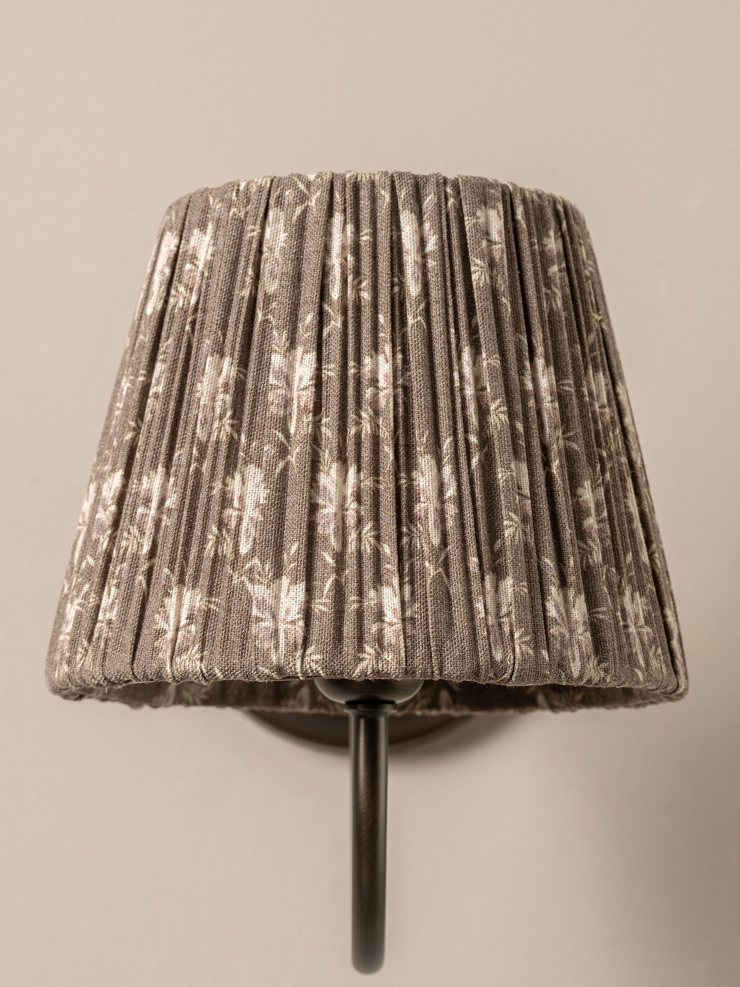 A pleated fabric lampshade in a taupe-brown and cream floral pattern, mounted on a dark metal wall sconce against a plain beige wall.