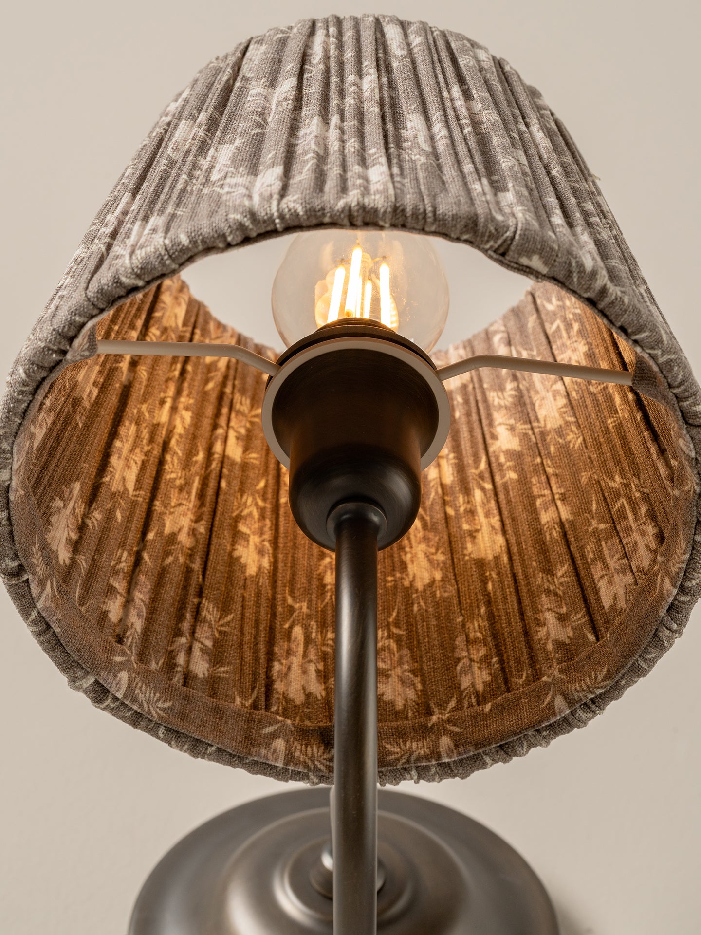 A close-up of a table lamp shade with a gray patterned outer fabric and a warm brown patterned inner shade, illuminated by a visible light bulb on a dark metal stand.