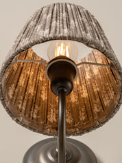 A close-up of a table lamp shade with a gray patterned outer fabric and a warm brown patterned inner shade, illuminated by a visible light bulb on a dark metal stand.