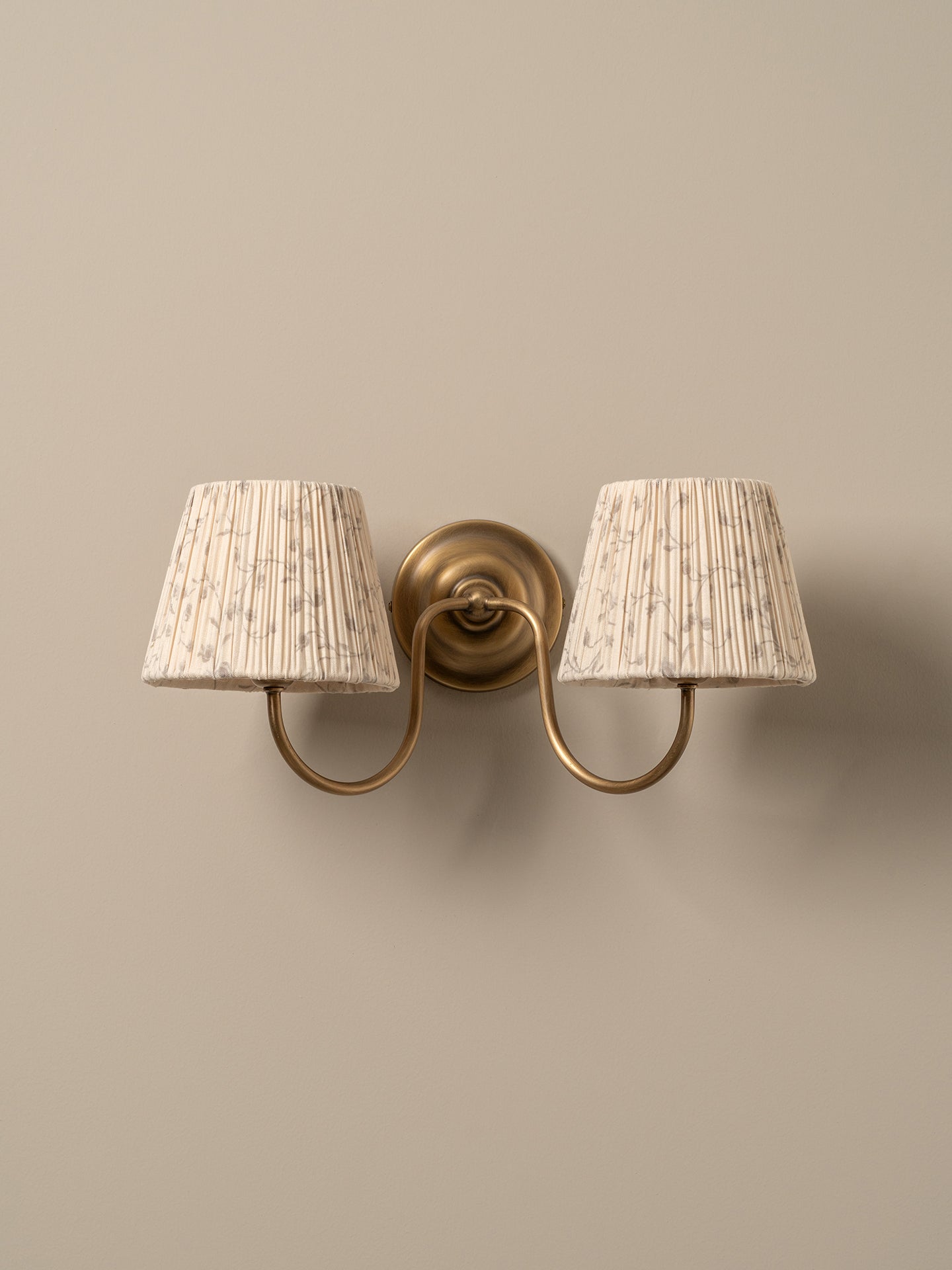 A two-light wall sconce with a warm brass-finished metal mount and curved arms, each holding a tapered pleated fabric shade in a light beige tone with subtle darker speckling, shown against a plain warm beige wall.