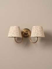 A two-light wall sconce with a warm brass-finished metal mount and curved arms, each holding a tapered pleated fabric shade in a light beige tone with subtle darker speckling, shown against a plain warm beige wall.