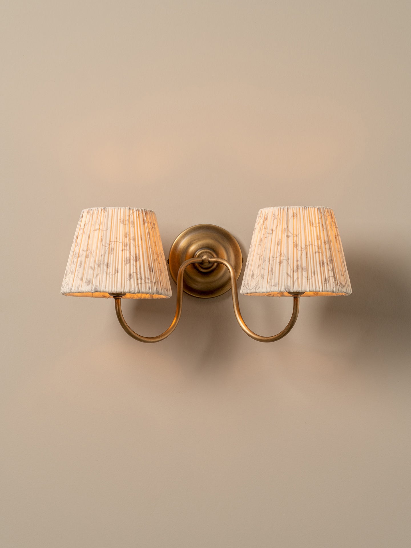 A two-light wall sconce with a warm brass-finished metal mount and curved arms, each holding a tapered off-white fabric shade with a subtle vertical textured pattern, shown against a plain beige wall.