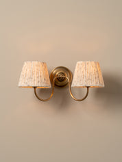 A two-light wall sconce with a warm brass-finished metal mount and curved arms, each holding a tapered off-white fabric shade with a subtle vertical textured pattern, shown against a plain beige wall.