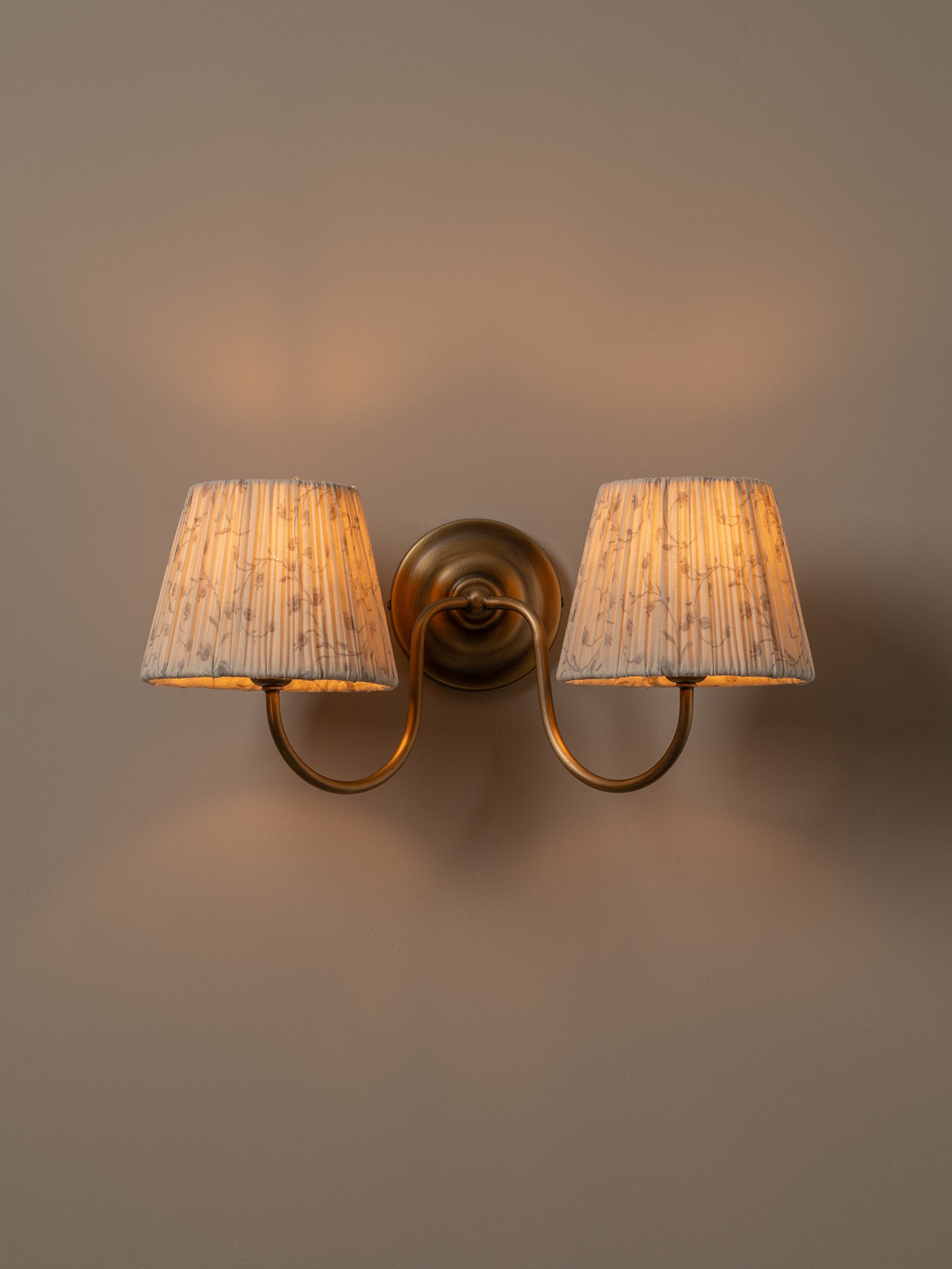 A two-light wall sconce with a warm brass-finished metal mount and curved arms, each holding a tapered beige fabric shade, shown lit against a plain taupe wall.