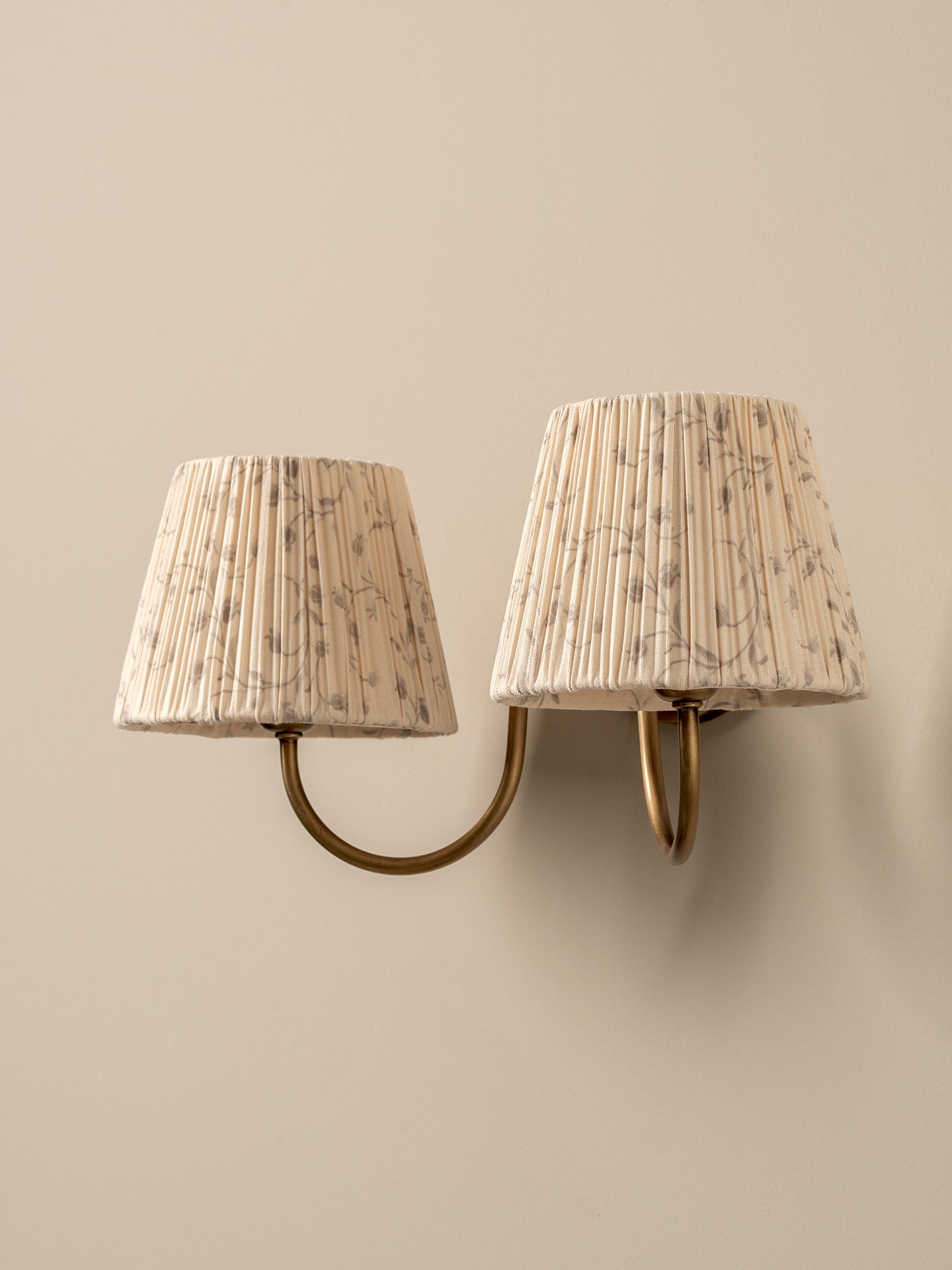 A two-light wall sconce with tapered pleated fabric shades in a beige and brown floral pattern, mounted on a warm beige wall.