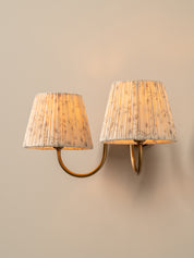 A two-light wall sconce with pleated beige and white patterned shades and curved gold metal arms, mounted on a warm beige wall.