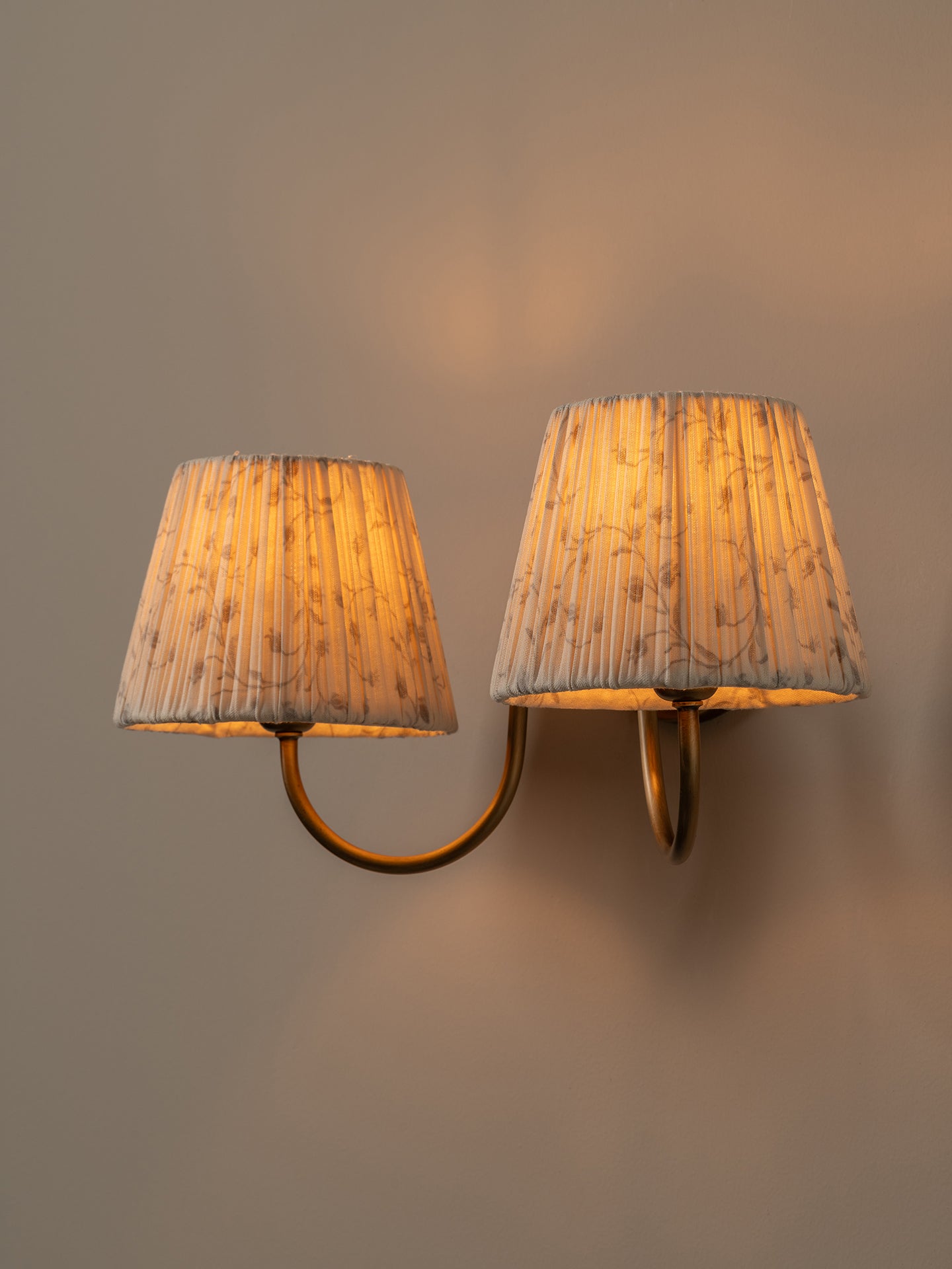 A double-arm wall sconce with pleated fabric shades, shown lit against a warm neutral wall.