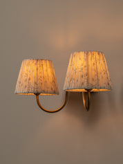 A double-arm wall sconce with pleated fabric shades, shown lit against a warm neutral wall.
