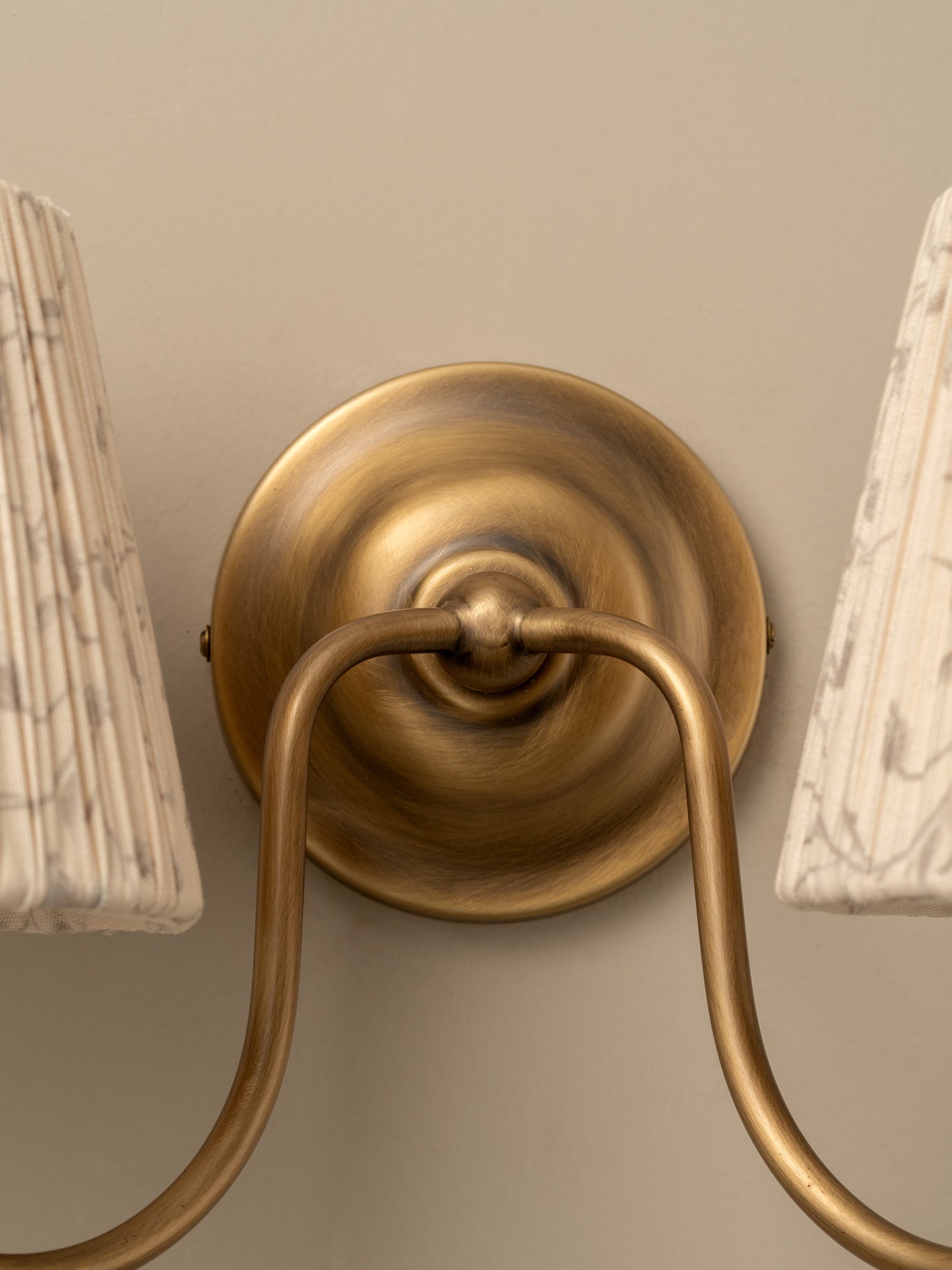 Close-up of a wall-mounted two-light sconce with an antique brass circular backplate and curved arms, flanked by two white pleated lampshades against a beige wall.