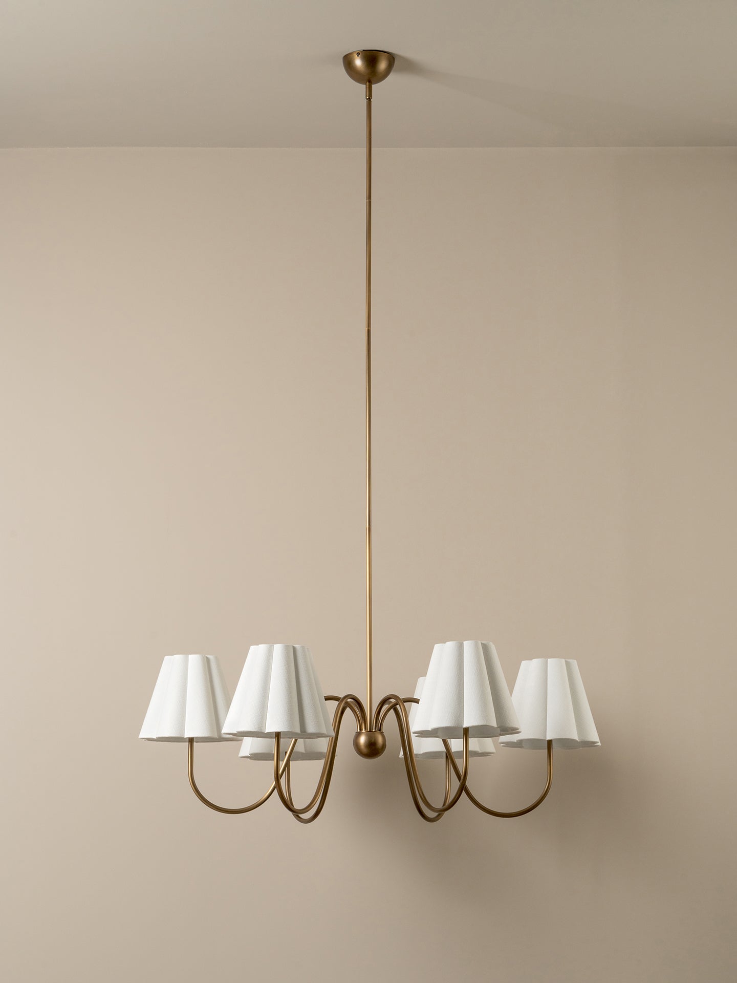 A modern six-light chandelier with a brushed brass or bronze metal frame and white tapered fabric shades, suspended from a matching ceiling canopy against a plain beige wall.
