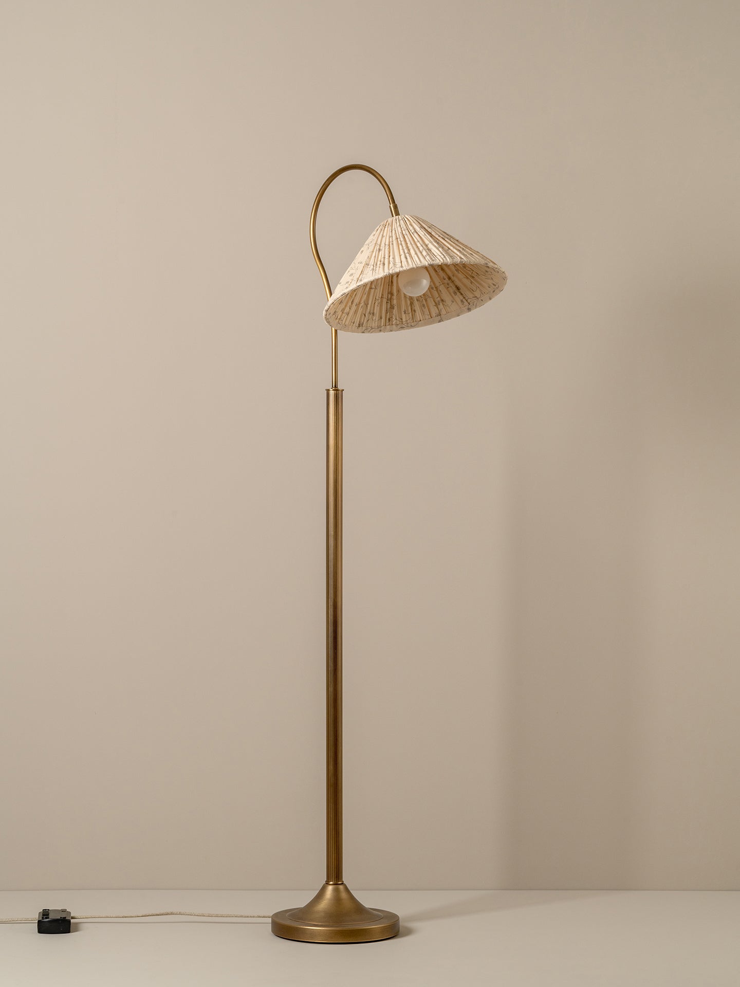 A vintage-style floor lamp with a warm brass finish and a conical shade made of woven natural fiber, glowing with a warm light against a neutral taupe background.