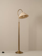 A vintage-style floor lamp with a warm brass finish and a conical shade made of woven natural fiber, glowing with a warm light against a neutral taupe background.