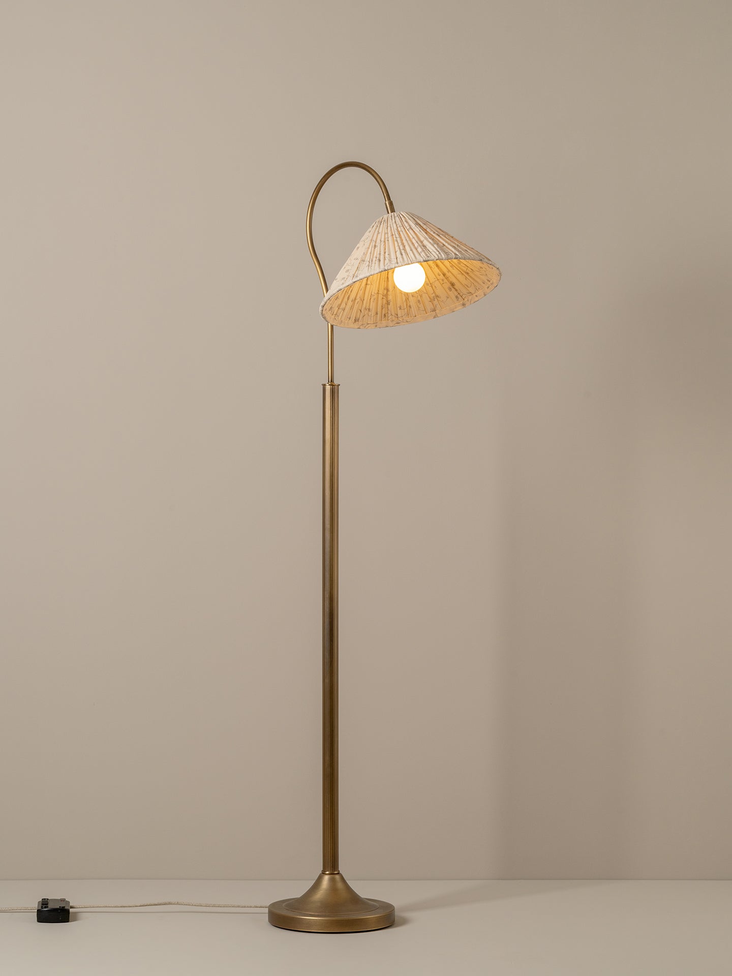 A vintage-style floor lamp with a warm brass finish and a conical shade made of woven natural fiber, glowing with a warm light against a neutral taupe background.