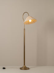A vintage-style floor lamp with a warm brass finish and a conical shade made of woven natural fiber, glowing with a warm light against a neutral taupe background.