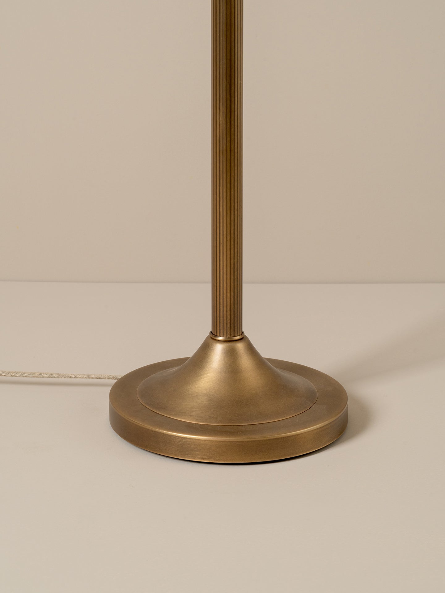 A vintage-style floor lamp with a warm brass finish and a conical shade made of woven natural fiber, glowing with a warm light against a neutral taupe background.