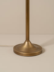 A vintage-style floor lamp with a warm brass finish and a conical shade made of woven natural fiber, glowing with a warm light against a neutral taupe background.
