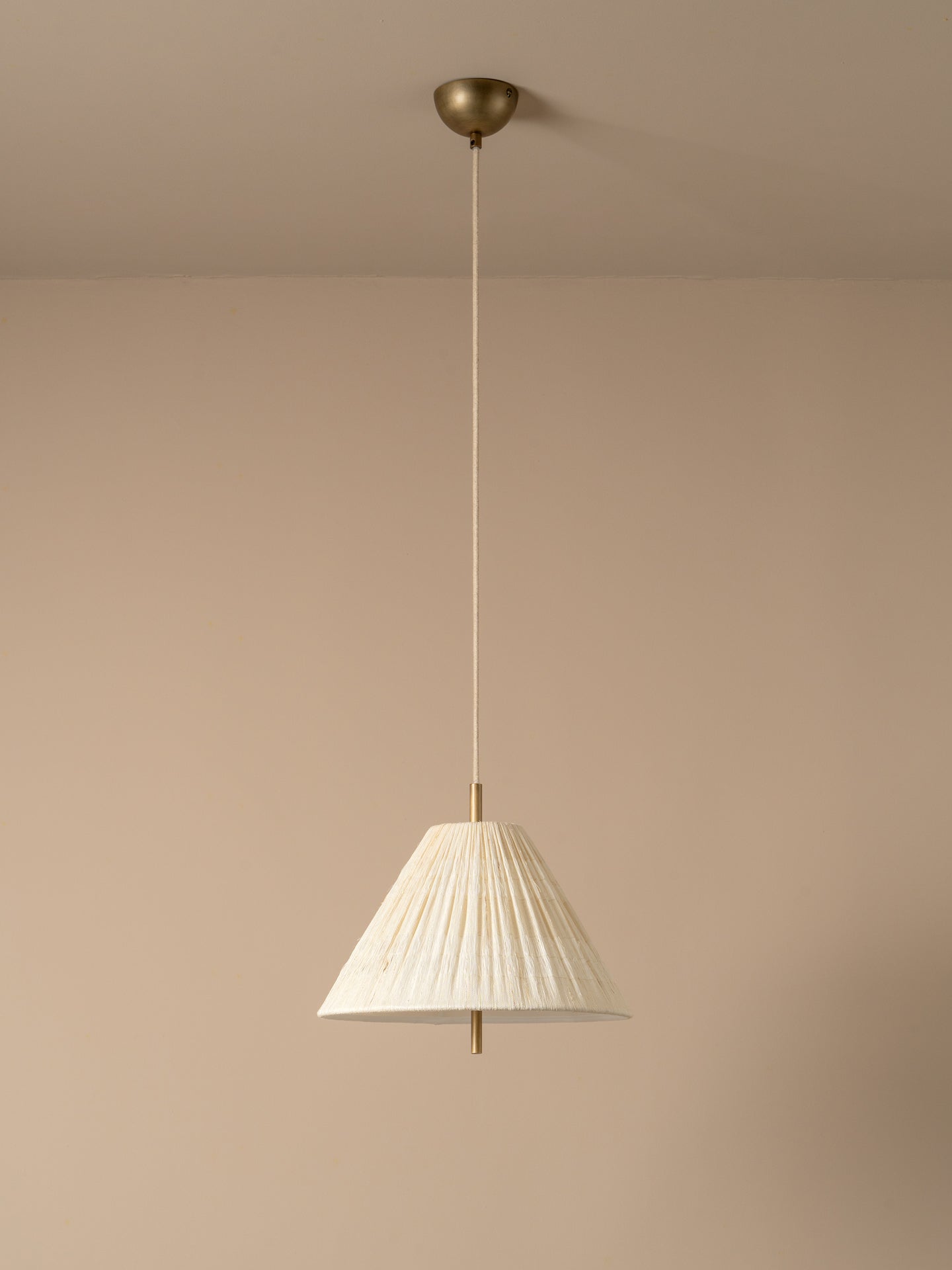 A modern pendant light with a pleated white fabric shade and a brass ceiling canopy, shown hanging against a minimalist beige wall and ceiling.